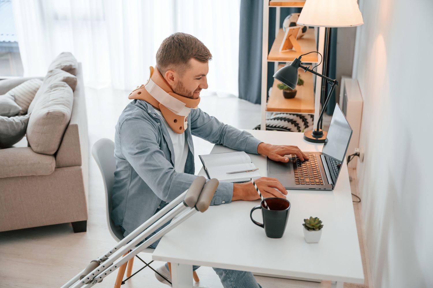 By the table with laptop. Man with crutches and neck brace is at home indoors.